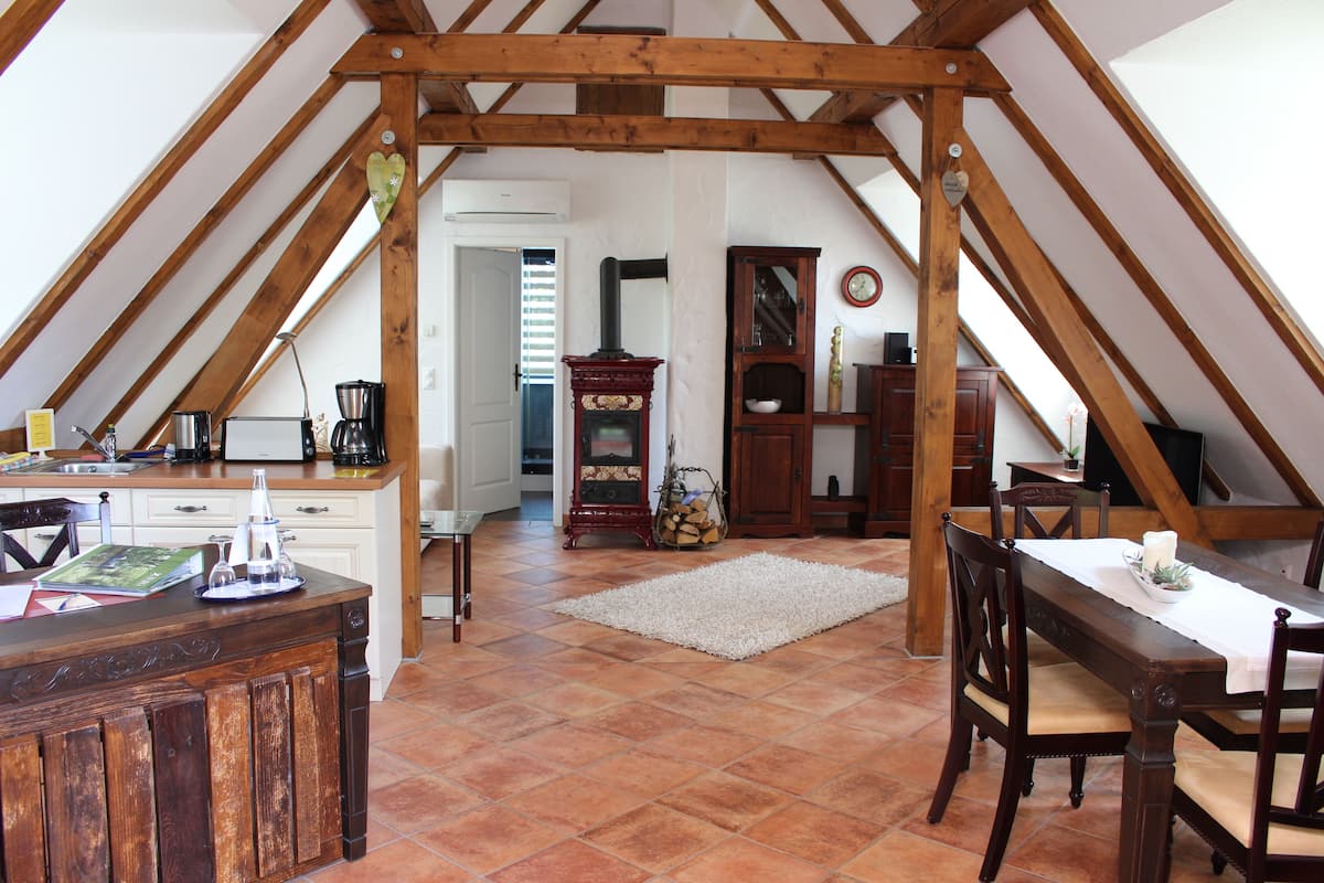 Una cocina rústica con vigas de madera, una zona de comedor con mesa y sillas, y suelo de baldosas.