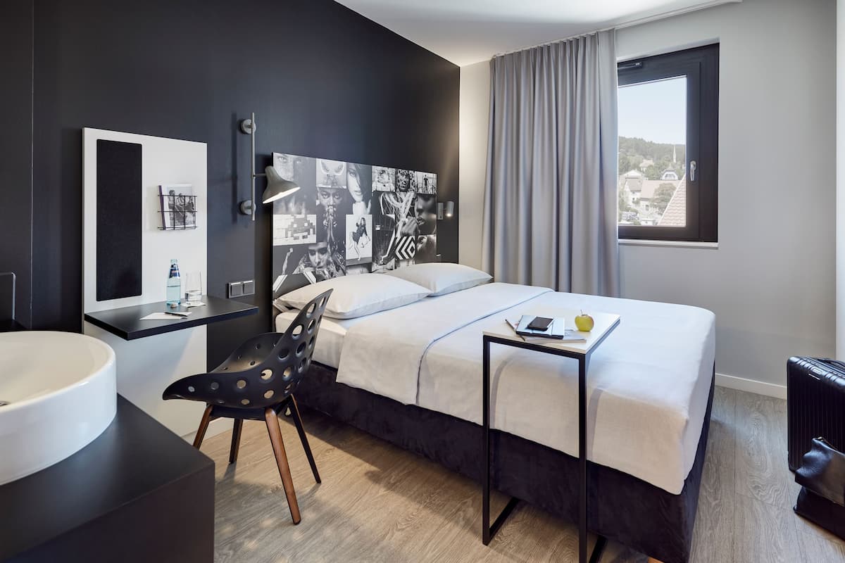 Quarto de hotel moderno com cama, mesa de cabeceira, cadeira e uma janela grande com vista.