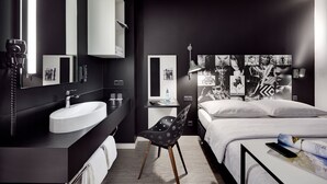 Una habitación de hotel moderna con una cama grande, un tocador blanco con lavabo, un escritorio con silla y una pared decorada con cuadros.