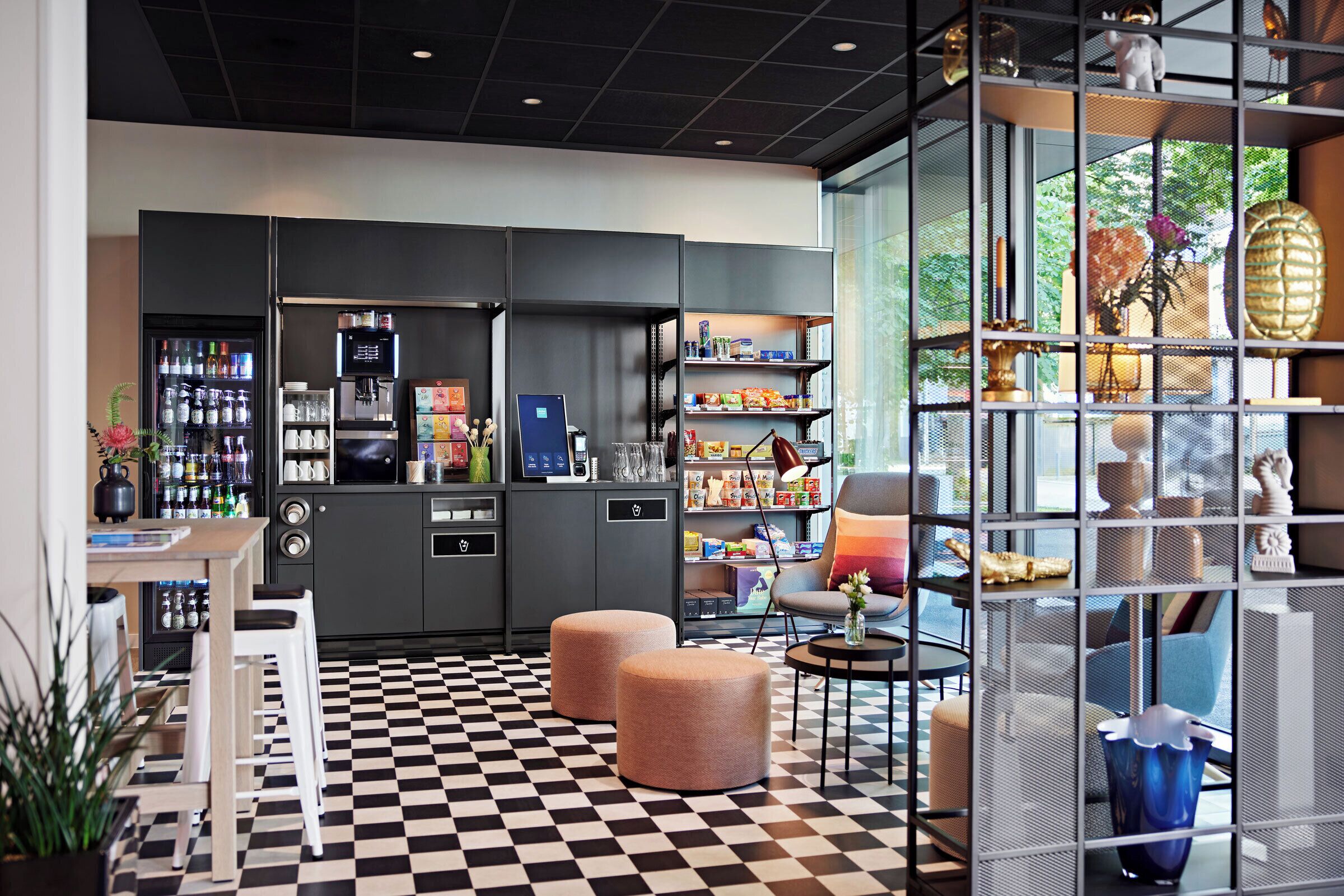 Un hall d’hôtel moderne avec un sol à carreaux, un espace salon, des distributeurs automatiques et une cloison en verre agrémentée d’objets décoratifs.