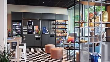 Un hall d’hôtel moderne avec un sol à carreaux, un espace salon, des distributeurs automatiques et une cloison en verre agrémentée d’objets décoratifs.