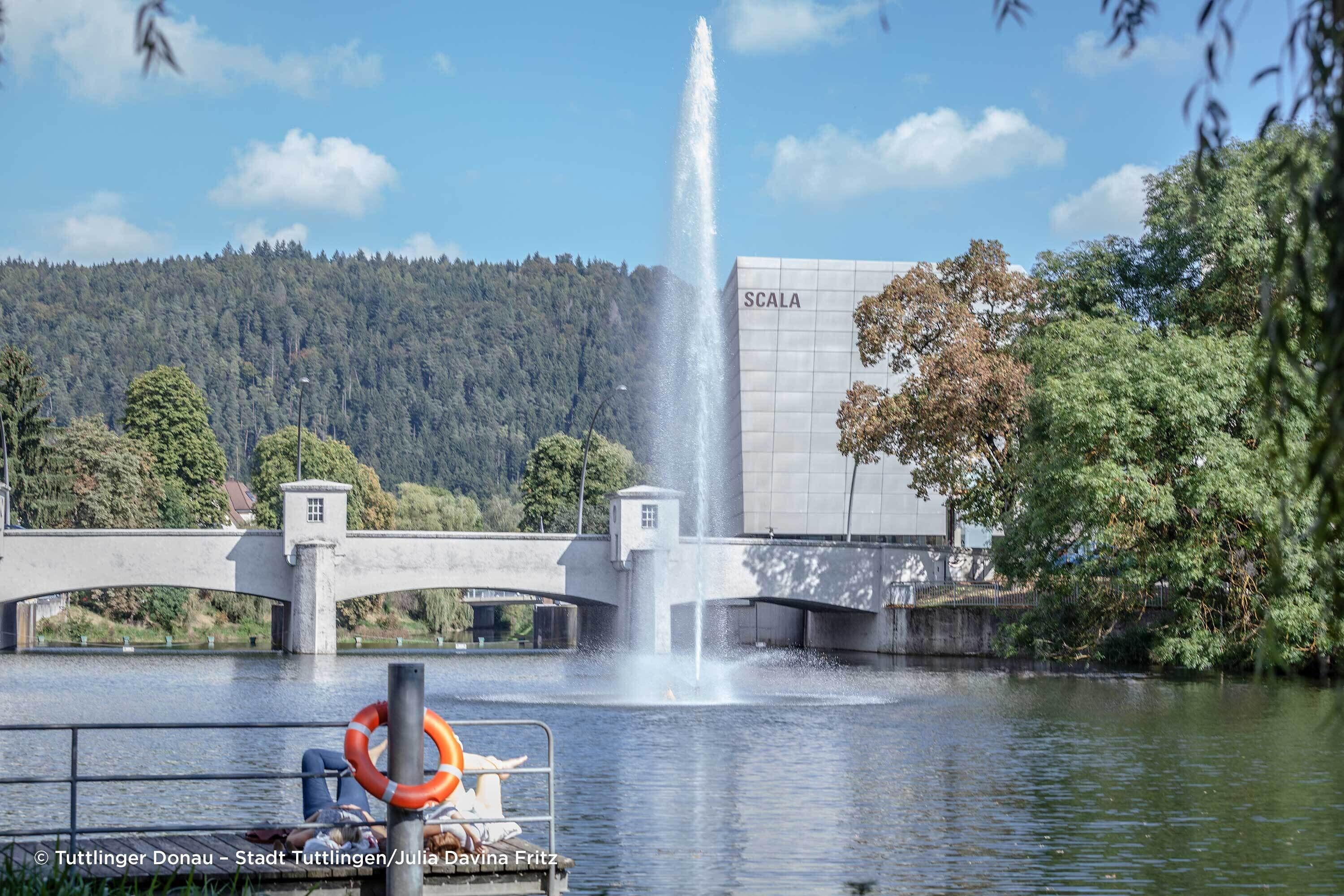 Un puente con una fuente, un flotador salvavidas y un edificio que dice "SCALA" cruzando un río.