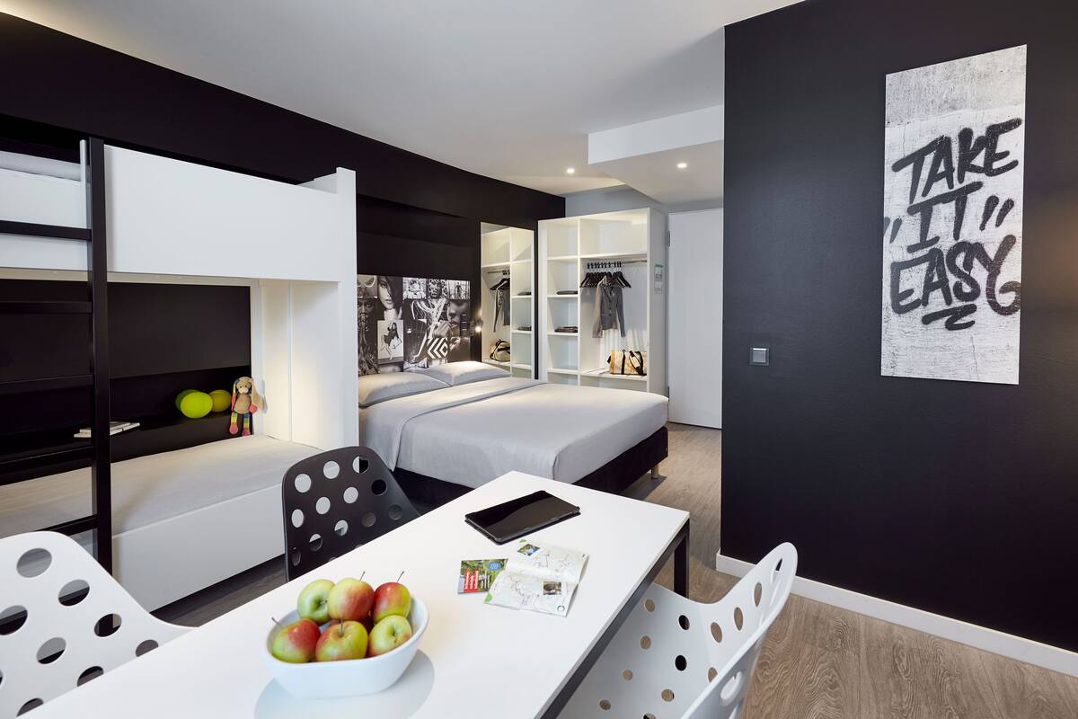 Um quarto moderno com cama, escrivaninha com tablet e uma tigela de frutas, guarda-roupa e um cartaz motivacional na parede.
