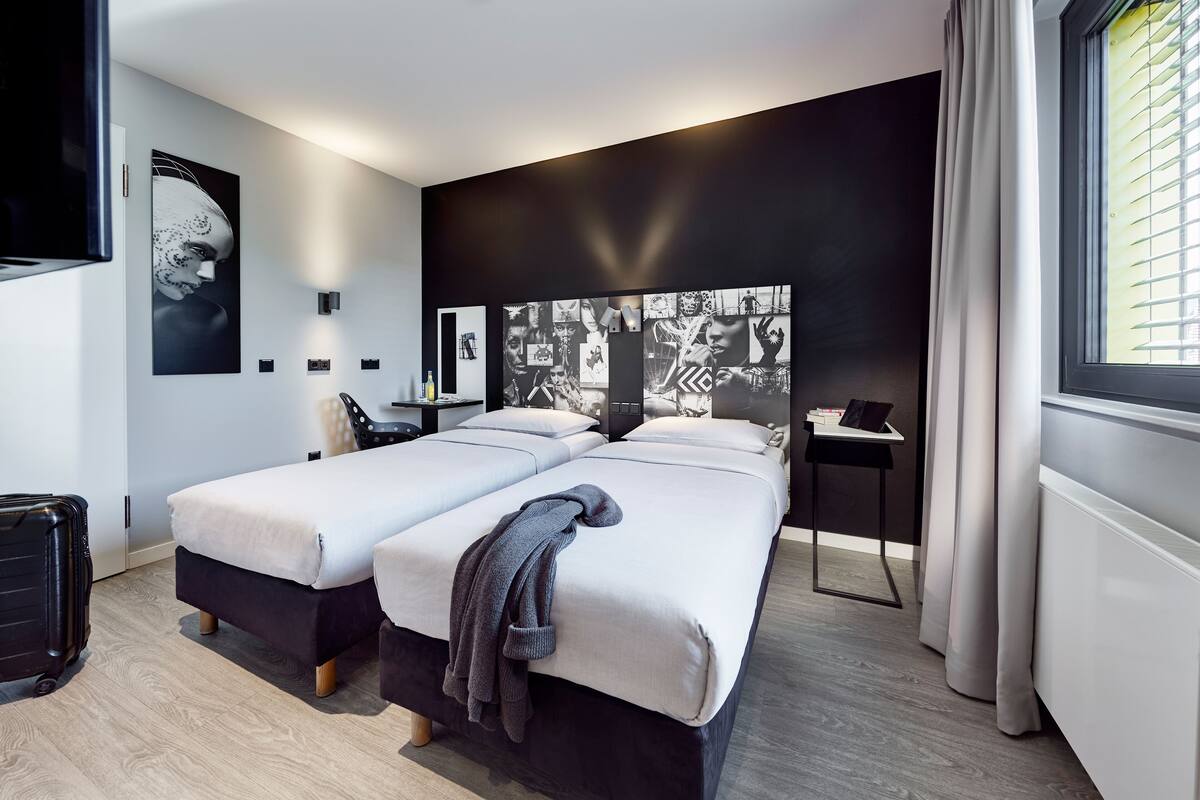 Quarto de hotel moderno com duas camas, uma grande obra de arte na parede, uma mesinha com abajur e uma janela com cortinas.