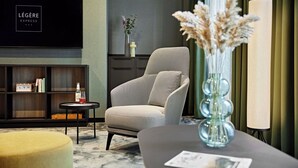 Una habitación de hotel moderna con un sillón gris, una mesita redonda pequeña con una botella, una estantería y un jarrón grande con flores secas.