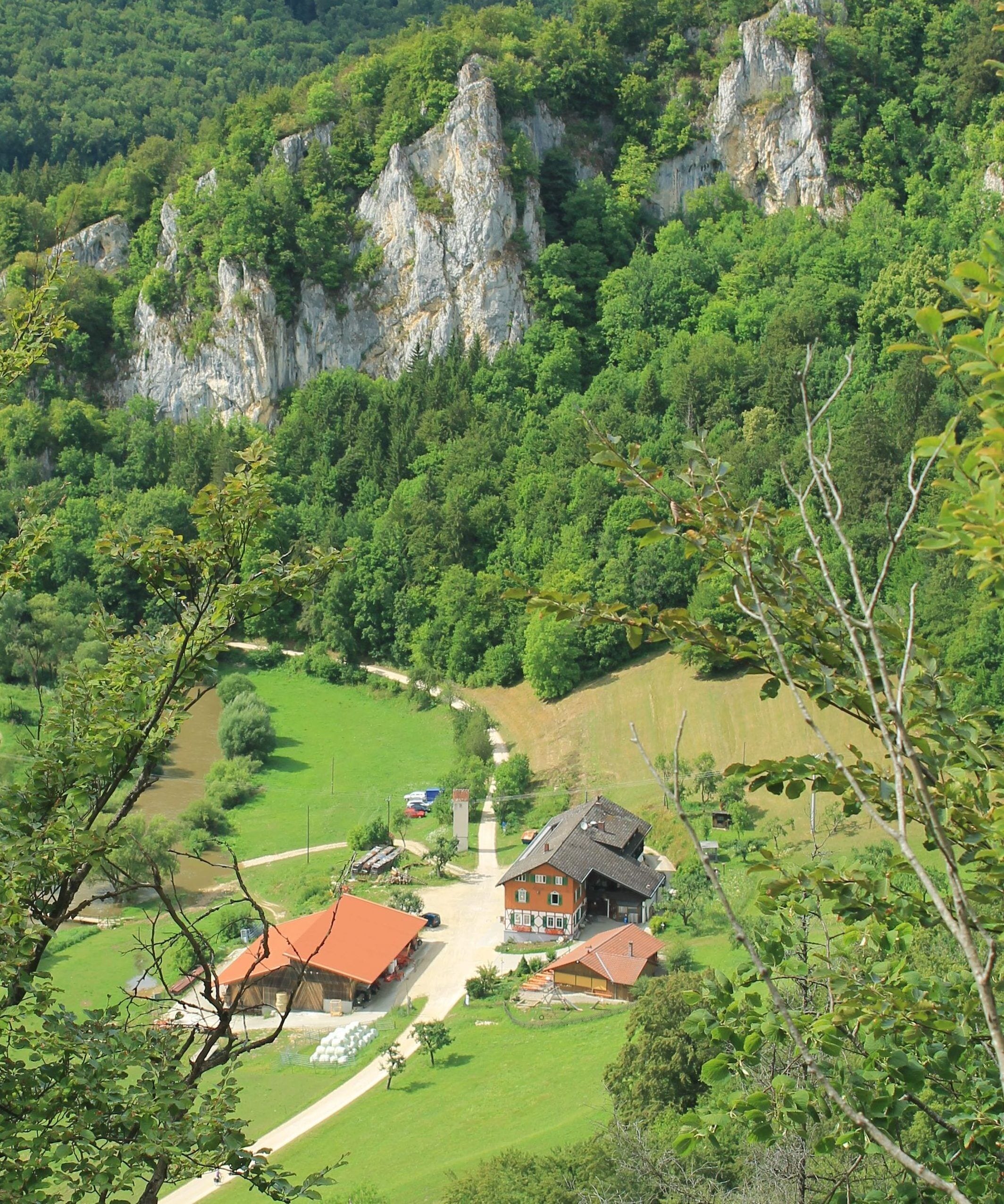 A rural house with a red roof, surrounded by lush greenery and a river.