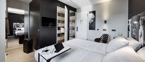 A modern hotel room with a large bed, a desk with a laptop, a chair, and a wardrobe.