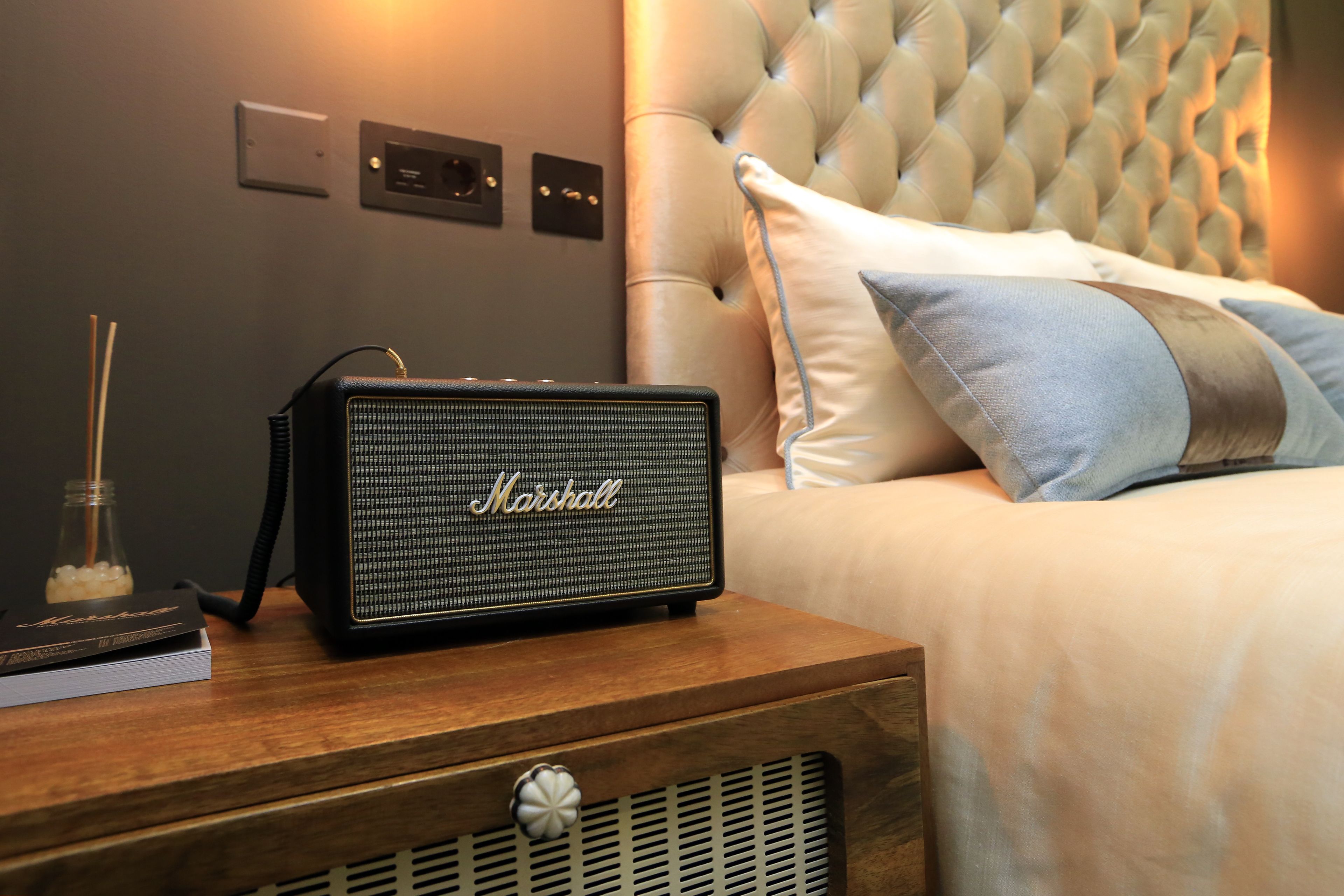 A Marshall speaker on a wooden nightstand next to a bed with a tufted headboard and a pillow.