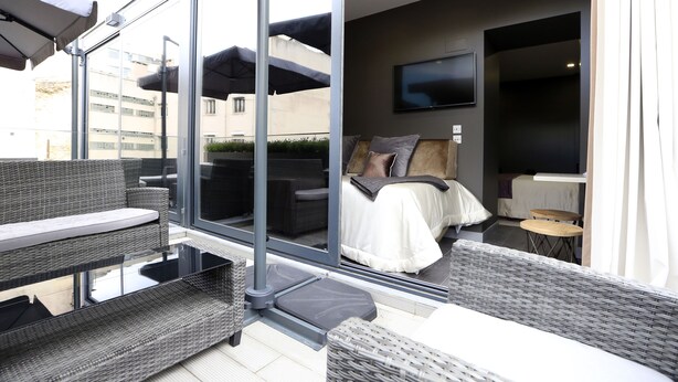 A modern balcony with wicker furniture, a glass door leading to a bedroom with a bed and a TV, and a small table.