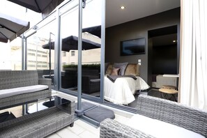 A modern balcony with wicker furniture, a glass door leading to a bedroom with a bed and a TV, and a small table. - Charm Flats (Lisbon)