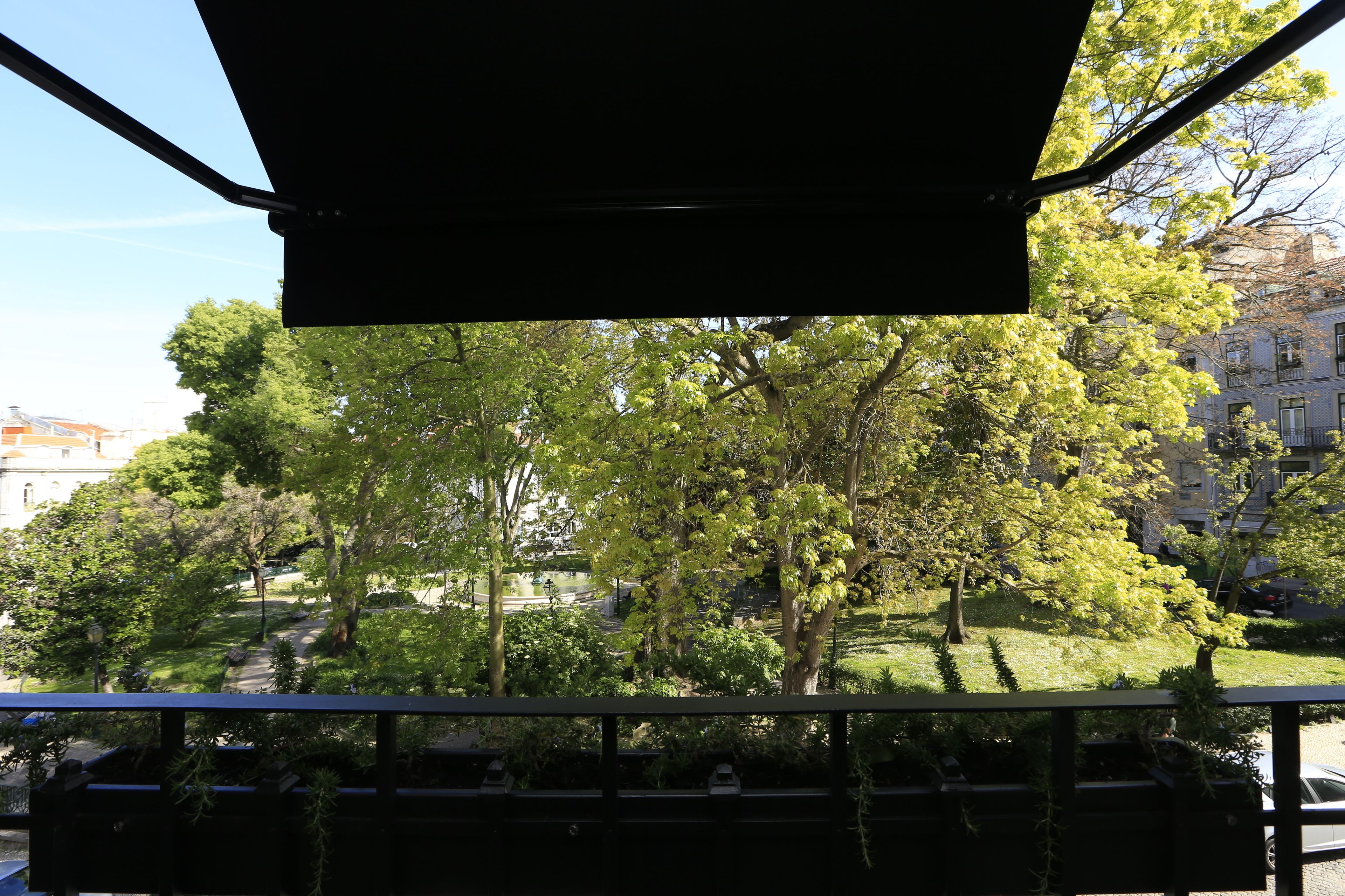A view from a balcony with a metal railing, overlooking a park with trees and a building in the background.