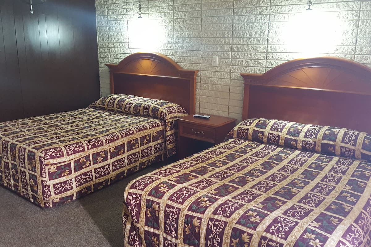 Camera d'albergo con due letti singoli, ognuno con testiera in legno e copripiumino decorato. Un comodino con un telecomando e pareti rivestite da carta da parati con texture.