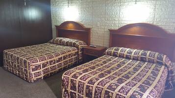 A hotel room with two single beds, each with a wooden headboard and a patterned bedspread. There is a nightstand with a remote control on it, and the walls are covered with a textured wallpaper.