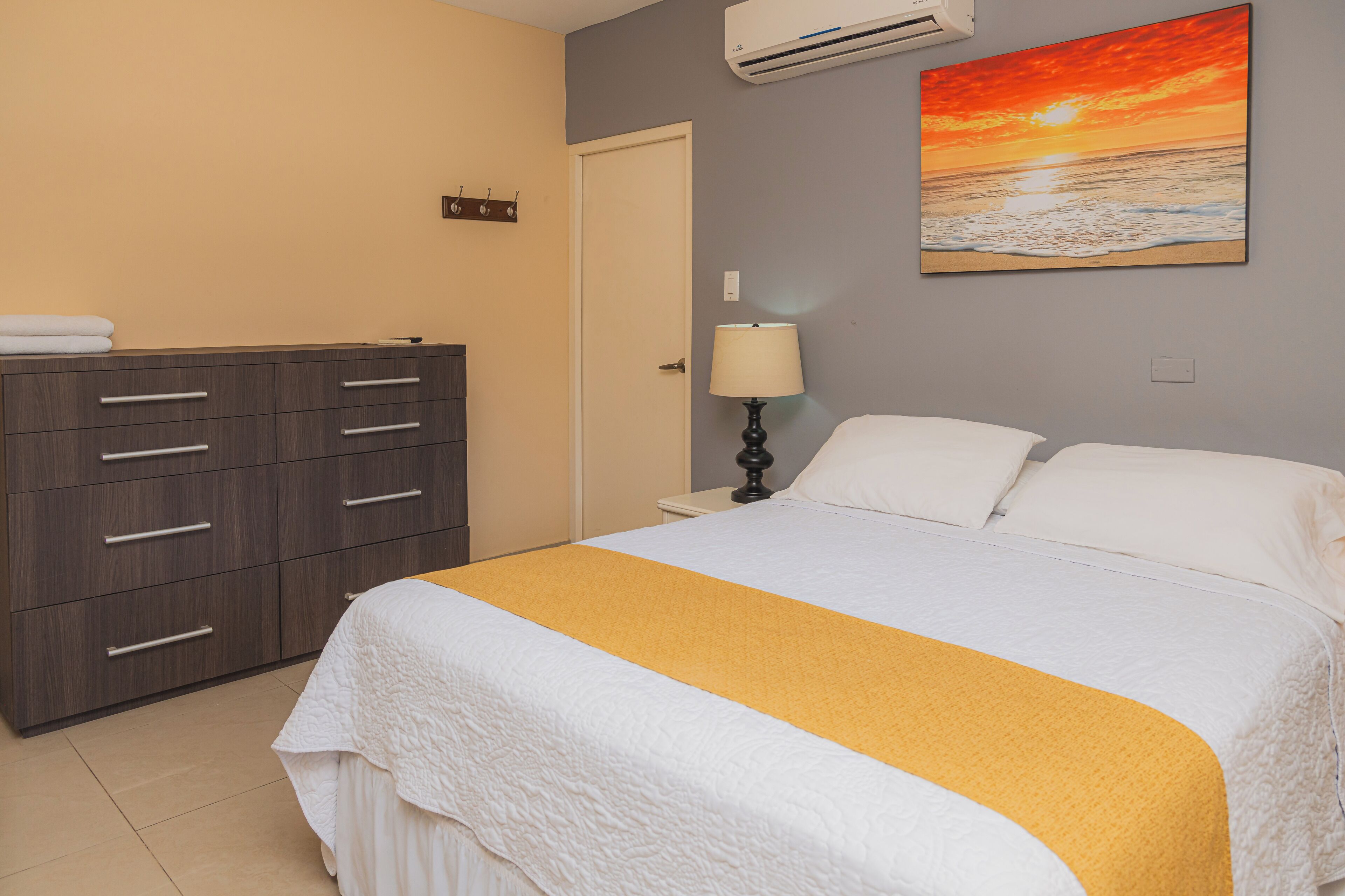A bedroom with a bed, a dresser, a nightstand, a lamp, and a painting of a sunset over the ocean.