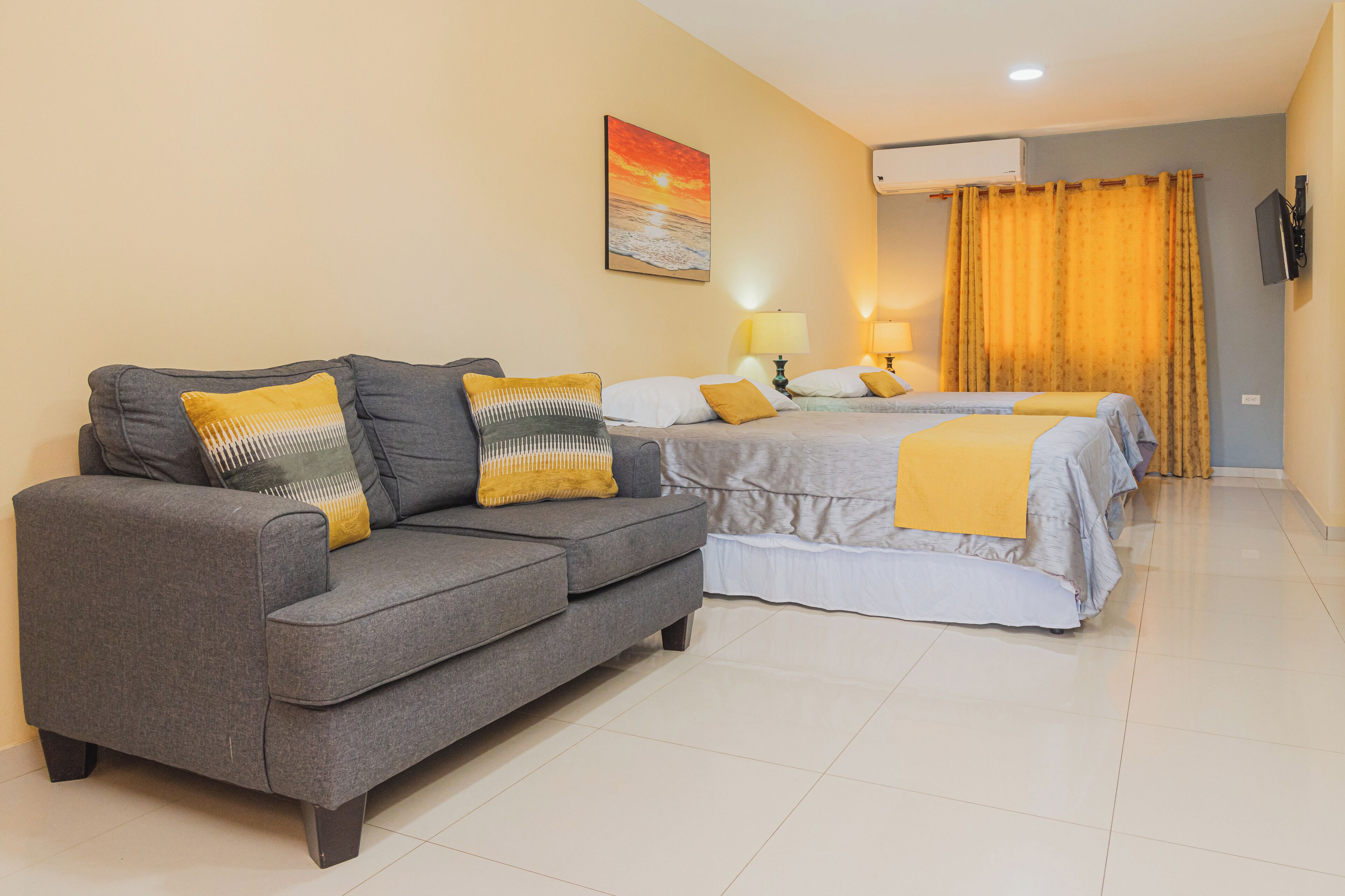 A hotel room with a grey sofa, two beds with yellow and white bedding, a painting of a sunset, and an air conditioning unit.