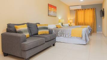 A hotel room with a grey sofa, two beds with yellow and white bedding, a painting of a sunset, and an air conditioning unit.