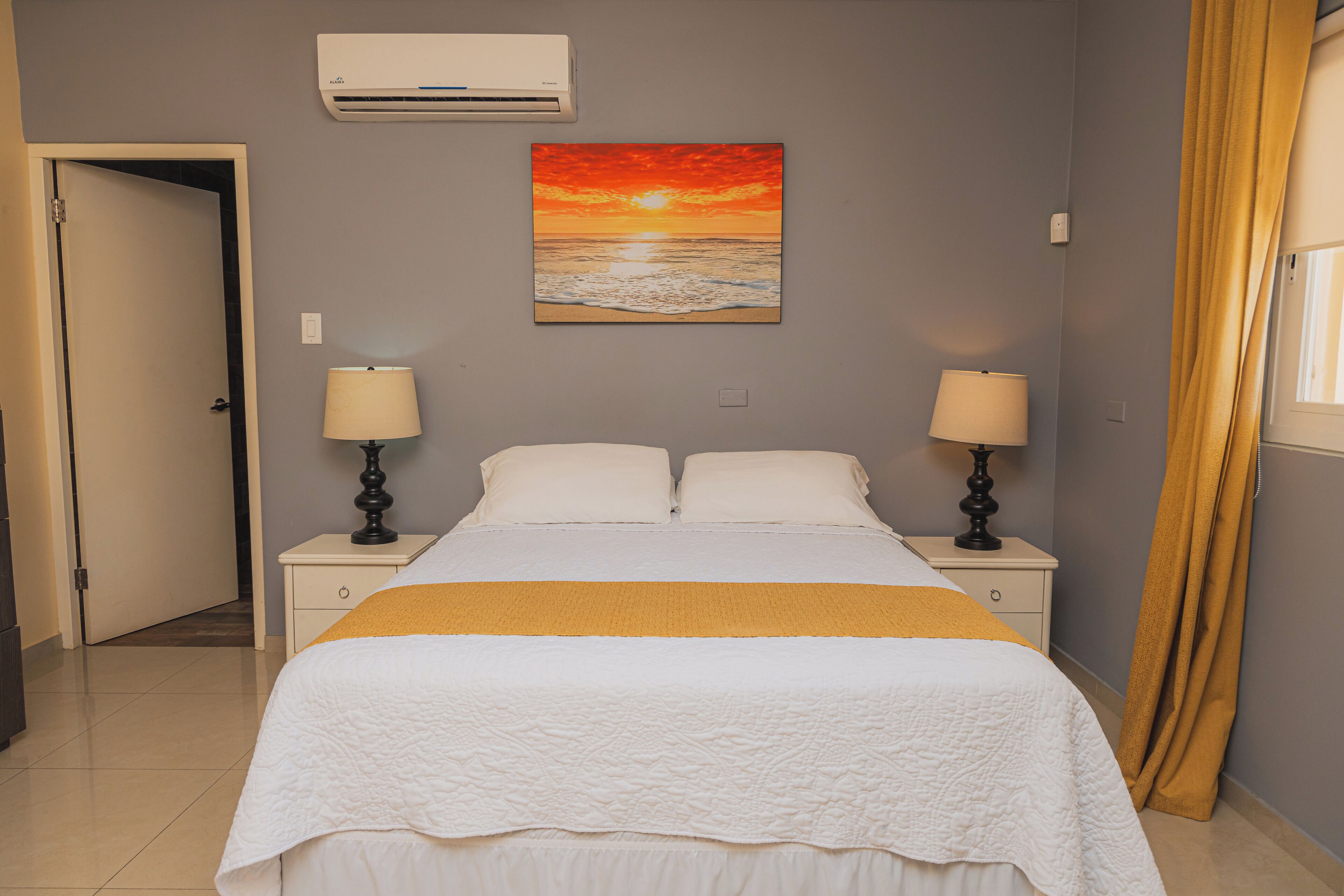 A bedroom with a bed, two bedside tables with lamps, a painting of a sunset, and a door.