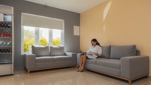 A living room with a grey sectional sofa, a coffee machine, and a window with a view of greenery. - Juanedu Suites (Oranjestad)