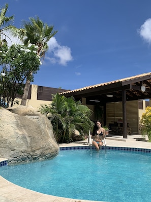 A pool area with a covered seating area, palm trees, and a large rock. - Juanedu Suites (Oranjestad)