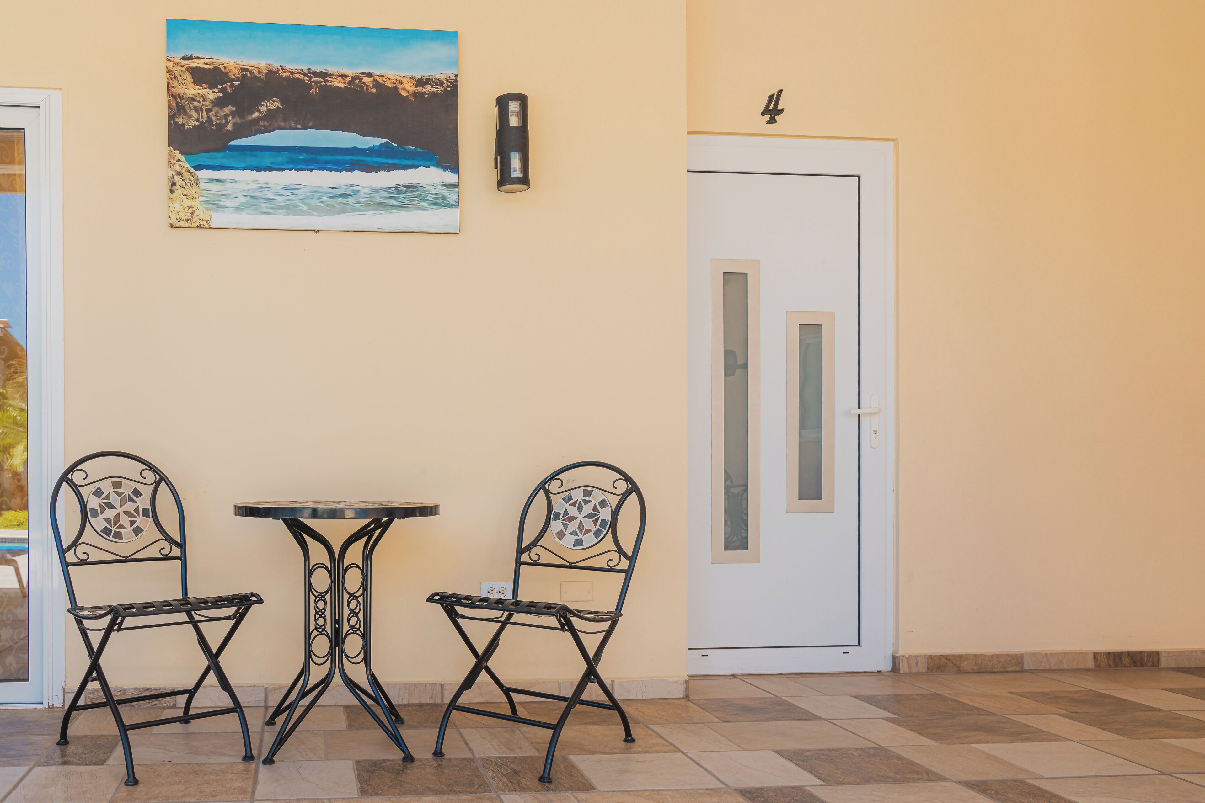 Una habitación con piso a cuadros, dos sillas de metal, una pequeña mesa redonda, una puerta con un número y un cuadro en la pared.