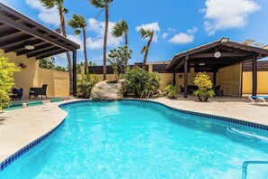 A swimming pool with a rock feature, surrounded by palm trees and lounge chairs. - Juanedu Suites (Oranjestad)