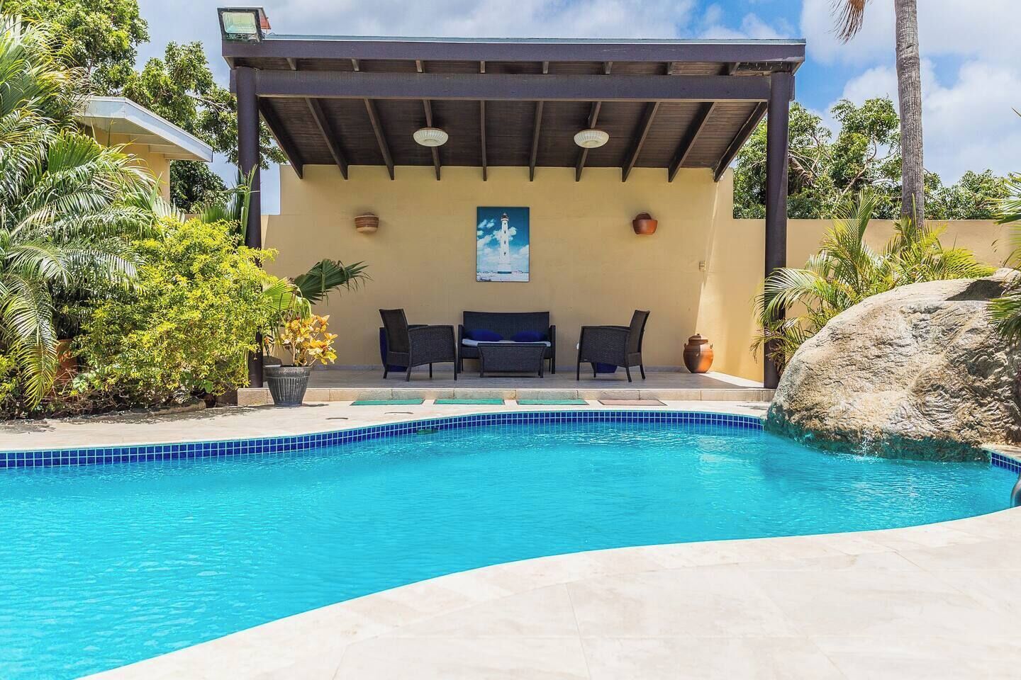 Una piscina con un área de estar techada, rodeada de plantas tropicales y una gran roca.