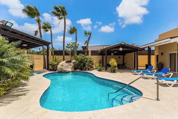 A swimming pool with a curved edge, surrounded by palm trees and lounge chairs. - Juanedu Suites (Oranjestad)