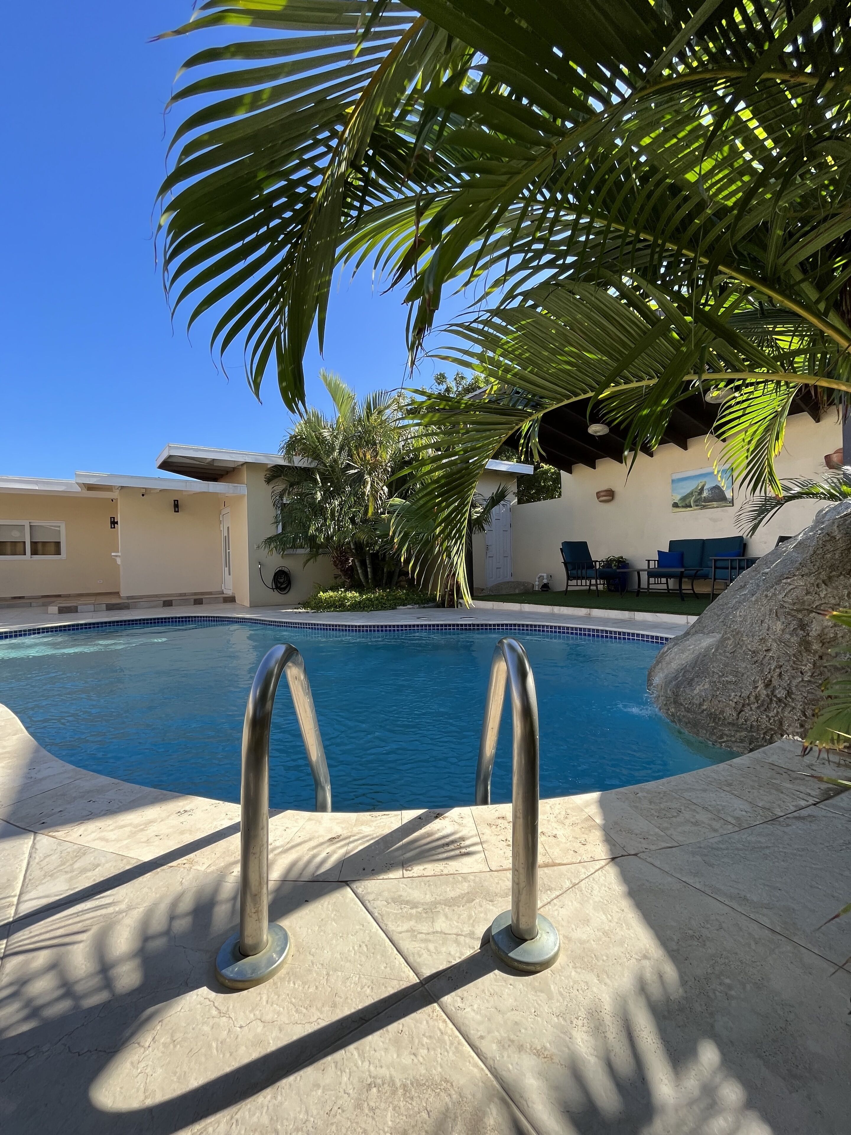 A swimming pool with metal ladders, surrounded by palm trees and a patio area with outdoor furniture.