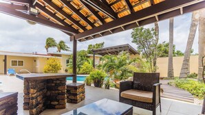 An outdoor patio area with stone and glass furniture, a pool, and lush landscaping. - Juanedu Suites (Oranjestad)