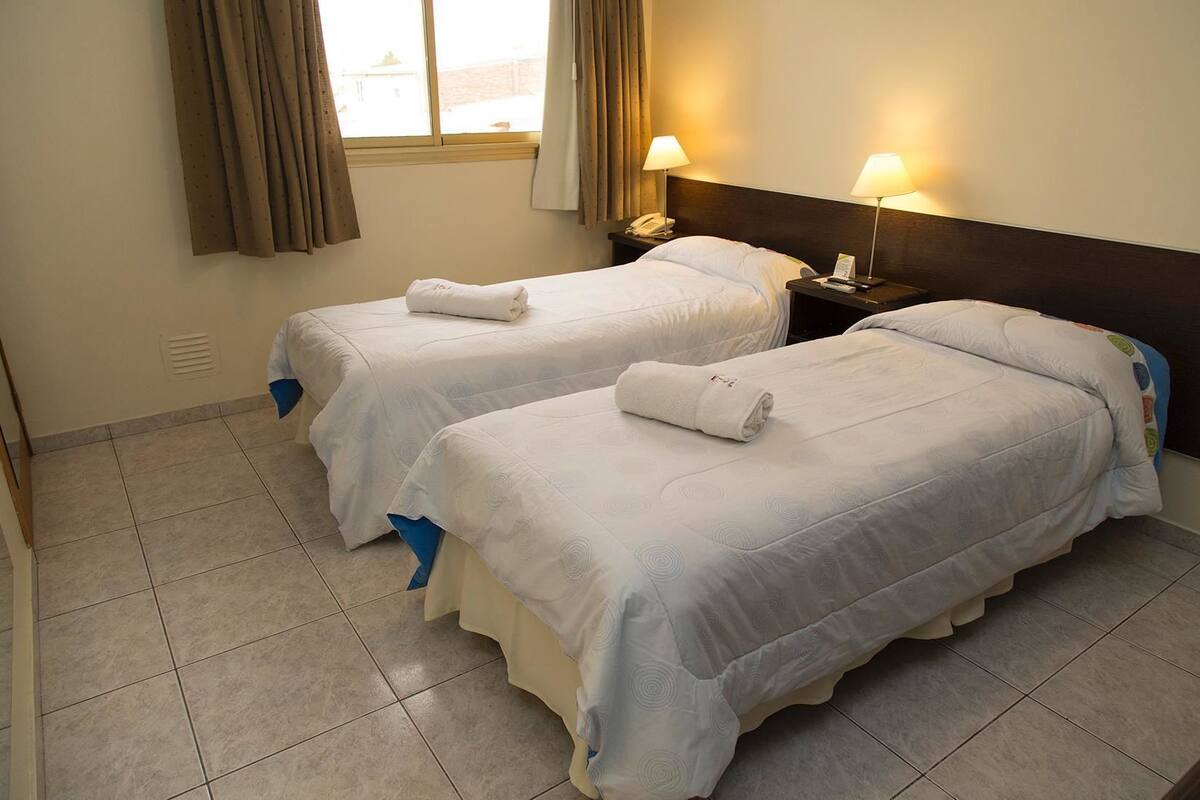 A hotel room with two single beds, each with a rolled towel, a nightstand with a lamp, and a window with curtains.