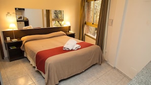 Habitación de hotel con cama, dos mesitas de noche, un espejo, ventana con cortinas y vista a árboles.