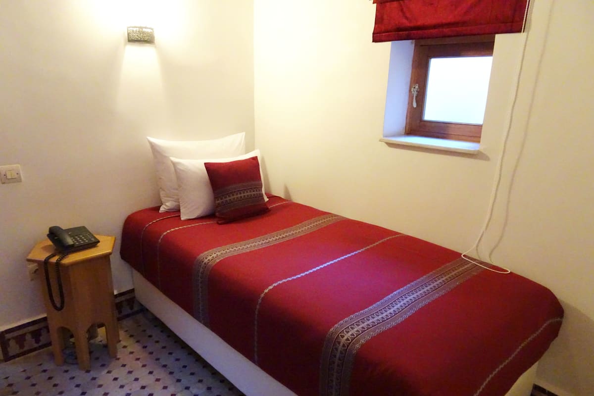 Una cama individual con una colcha roja y dos almohadas, un teléfono en la mesita de noche, un taburete de madera pequeño, una ventana con un visillo rojo y un suelo estampado.