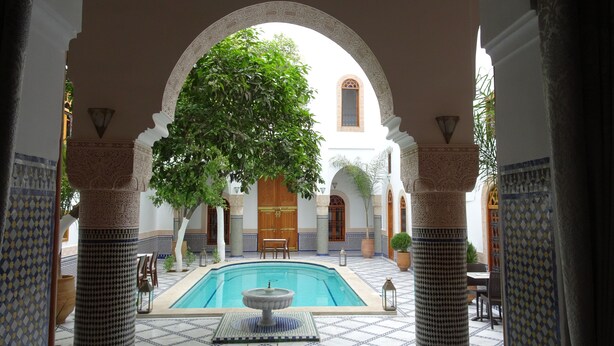Piscina interna com fonte, rodeada por colunas e arcos ornamentados, e vista para um edifício com porta de madeira.