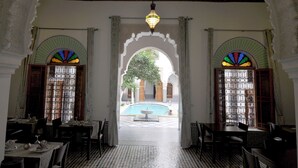 Uma sala de jantar com mesas e cadeiras, teto ornamentado e vista para um pátio com piscina.