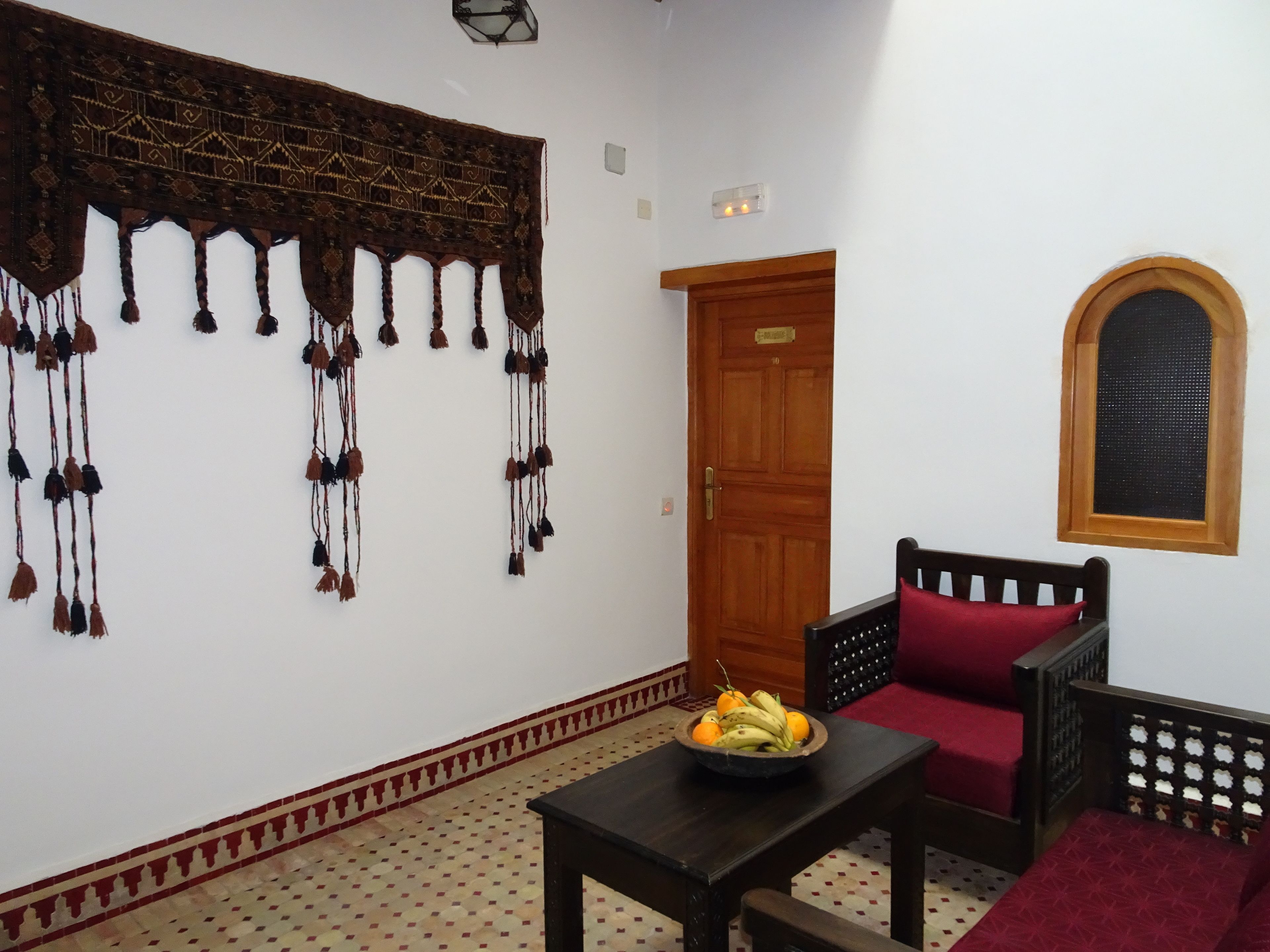 Una habitación con un tapiz decorativo en la pared, una puerta de madera, una pequeña mesa con frutero, una silla con cojín rojo y una ventana con marco de madera.
