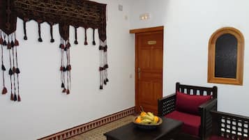 Una habitación con un tapiz decorativo en la pared, una puerta de madera, una pequeña mesa con frutero, una silla con cojín rojo y una ventana con marco de madera.