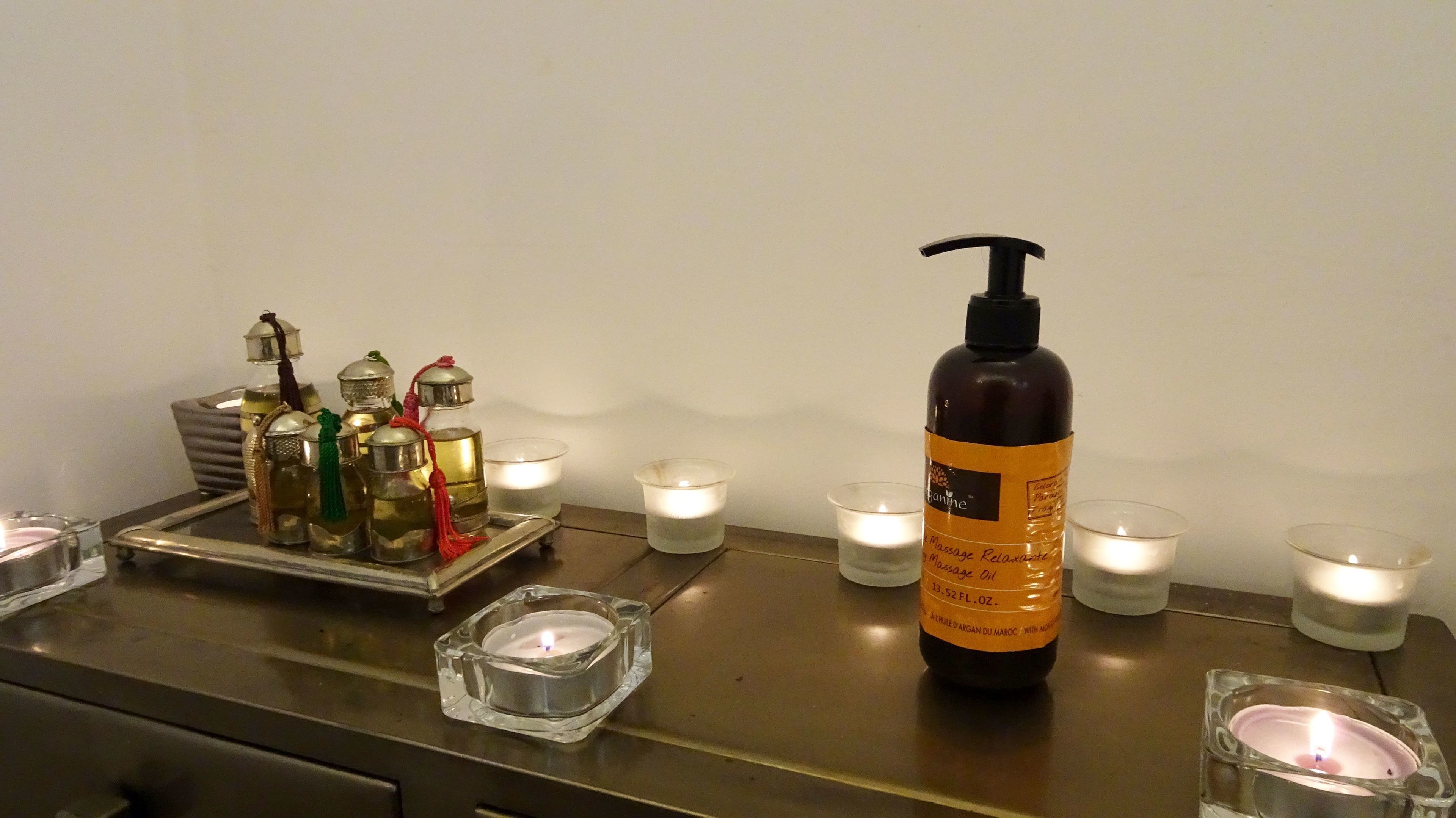 A bathroom counter with a bottle of massage oil, a tray with various bottles, and several lit candles.