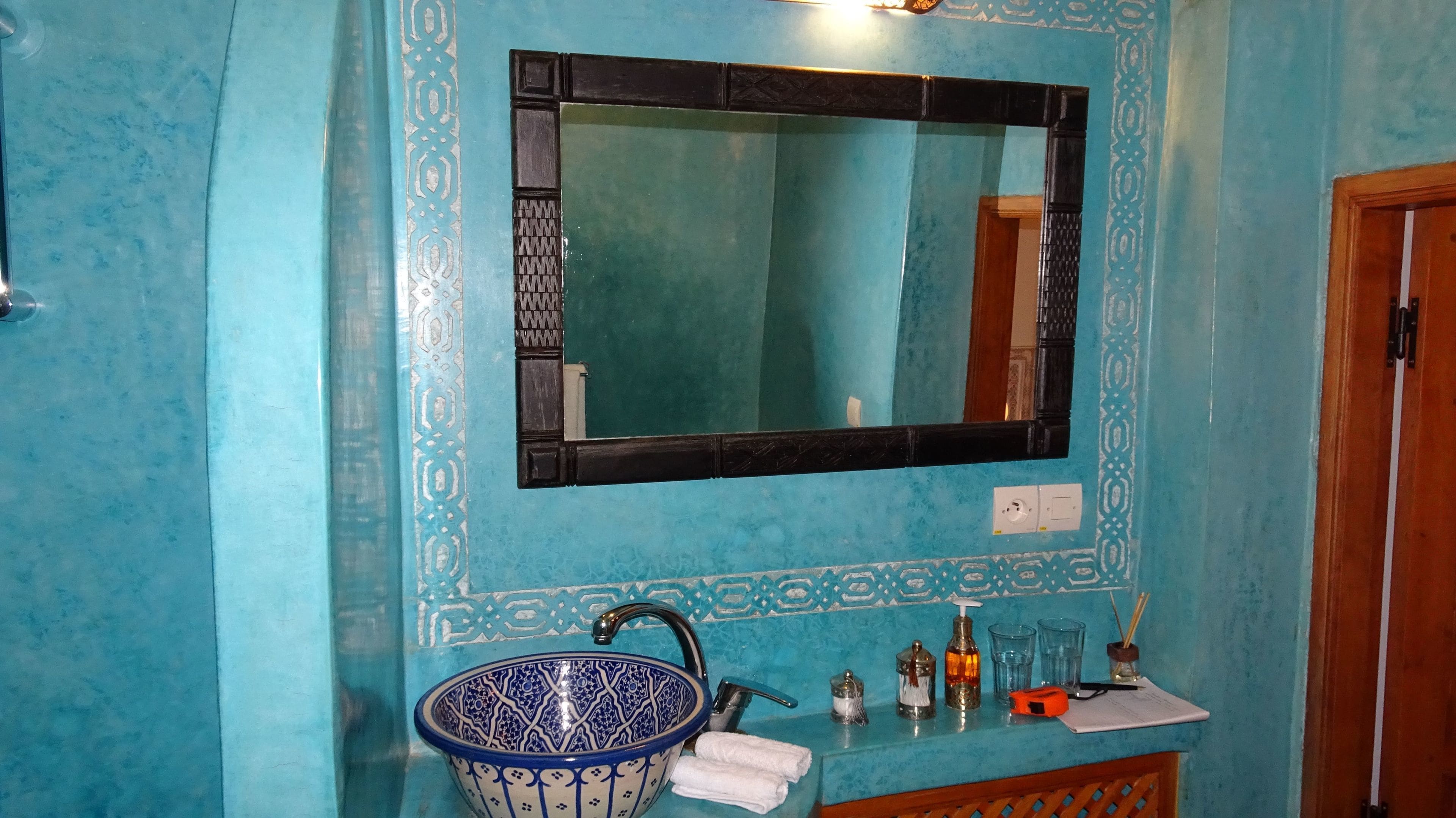 A bathroom with a blue tiled wall, a sink with a blue and white bowl, a mirror, and a wooden door.