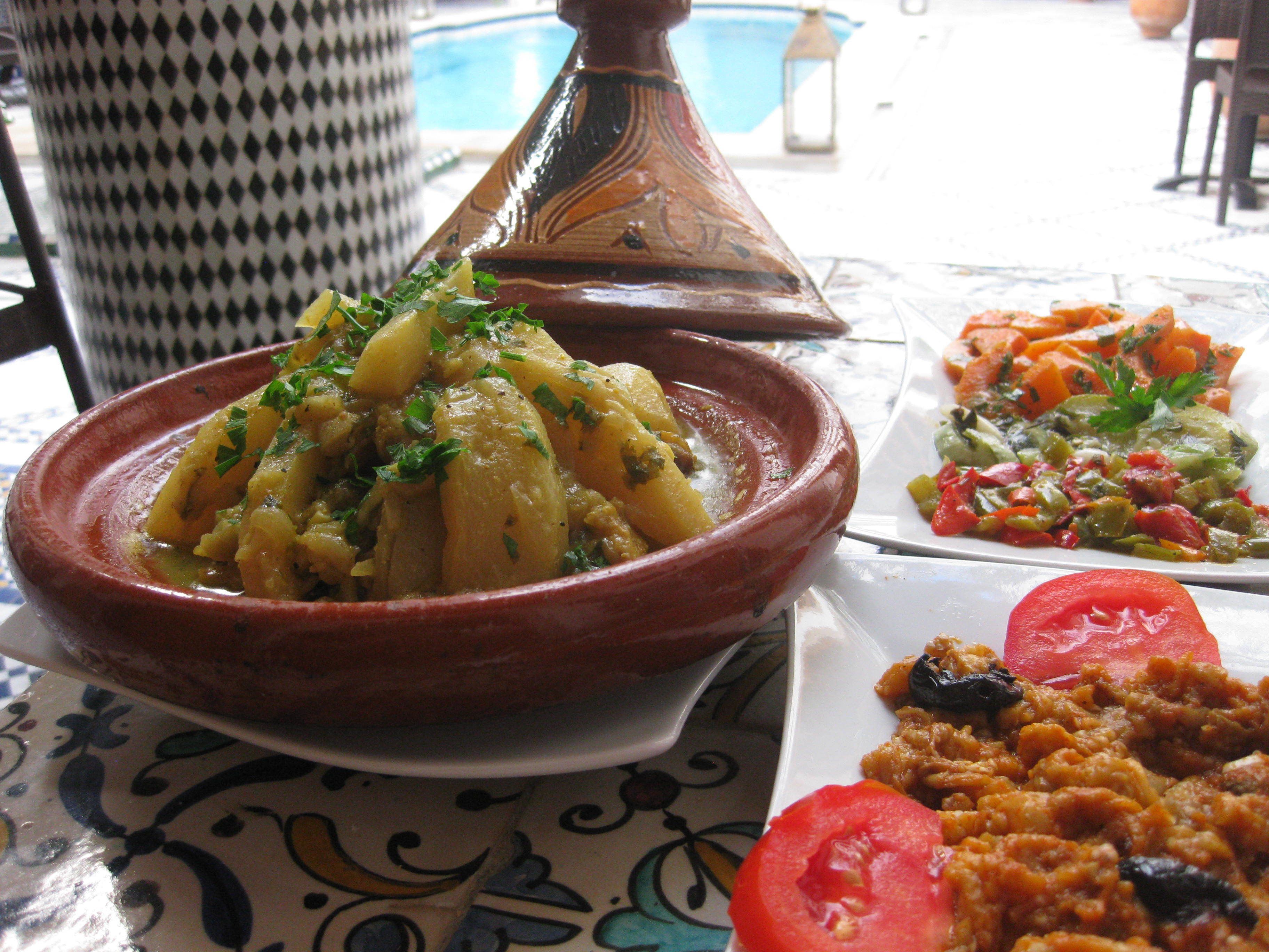 Uma mesa de jantar com um tagine marroquino, uma salada de legumes e um prato com tomates e passas.
