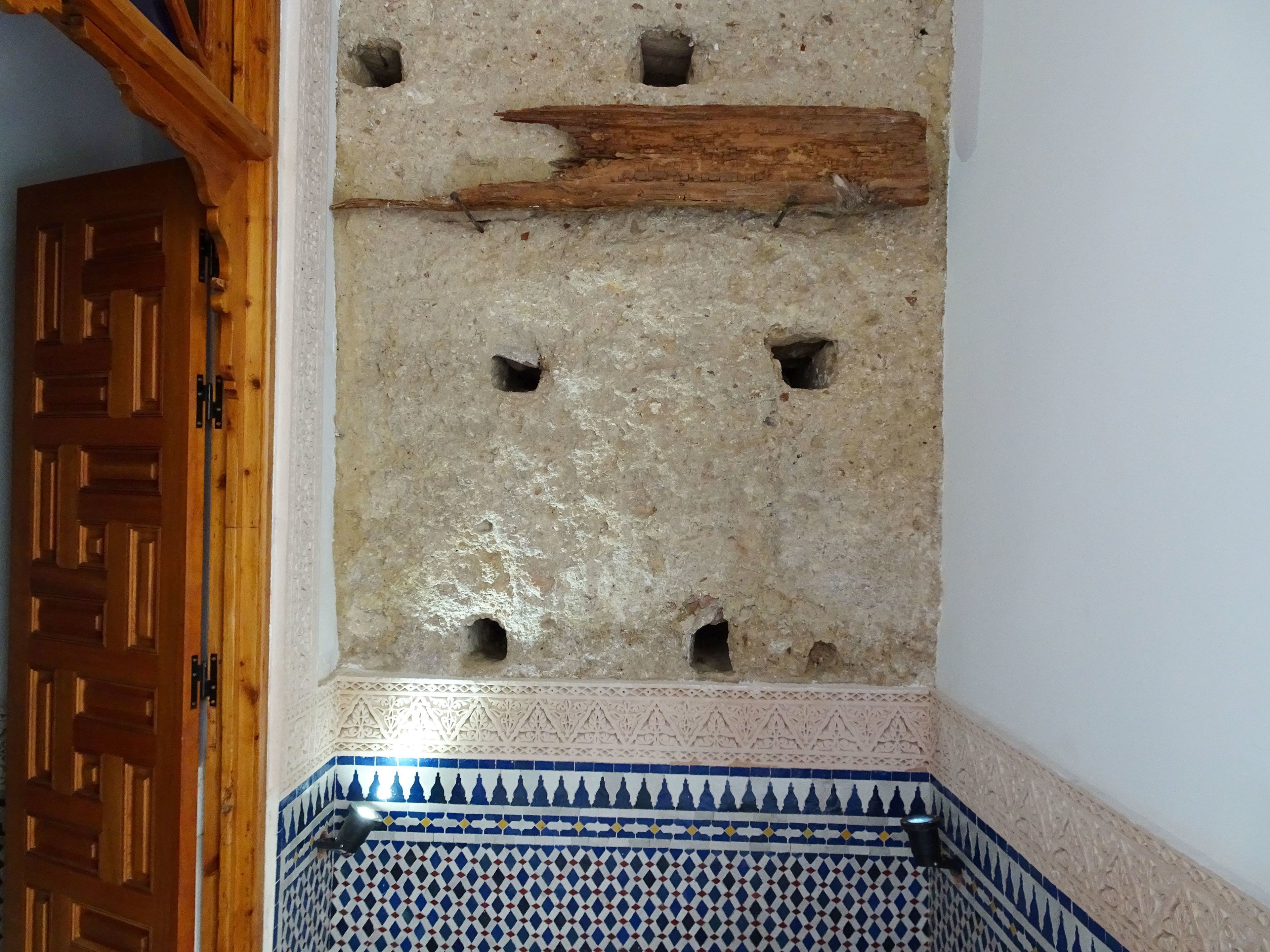Una habitación con pared de piedra que presenta varios agujeros, una puerta de madera y un borde de azulejos con diseño.
