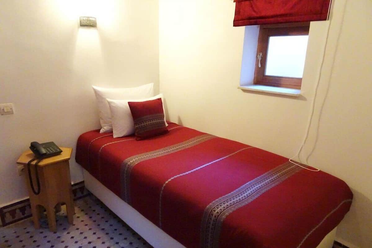 Una habitación pequeña con cama individual, colcha roja, teléfono en la mesita de noche, un taburete pequeño y una ventana con cortinilla roja.