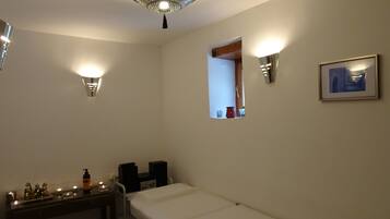 Quarto de hotel com cama, mesa de cabeceira com velas, uma pequena janela e luminárias na parede.
