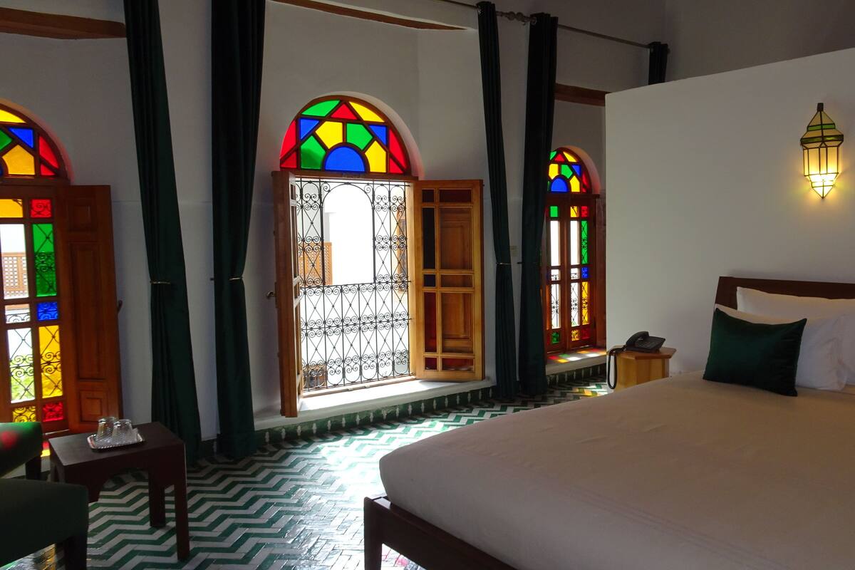 Una habitación de hotel con una cama grande, dos ventanas con vidrieras de colores, una mesita de noche con teléfono y una lámpara fijada a la pared.