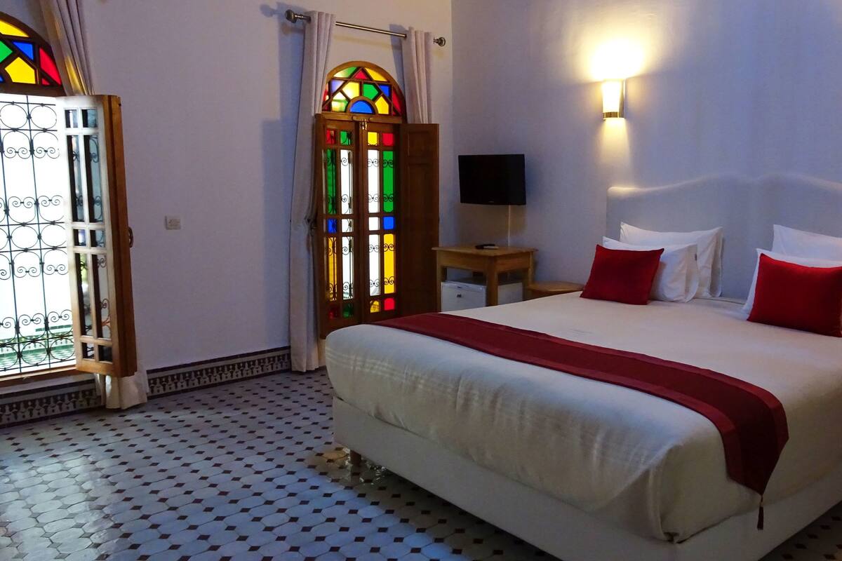 Una habitación de hotel con cama, televisor, mesita de noche, colcha roja y blanca, manta roja y una ventana con vitrales, además de un suelo estampado.
