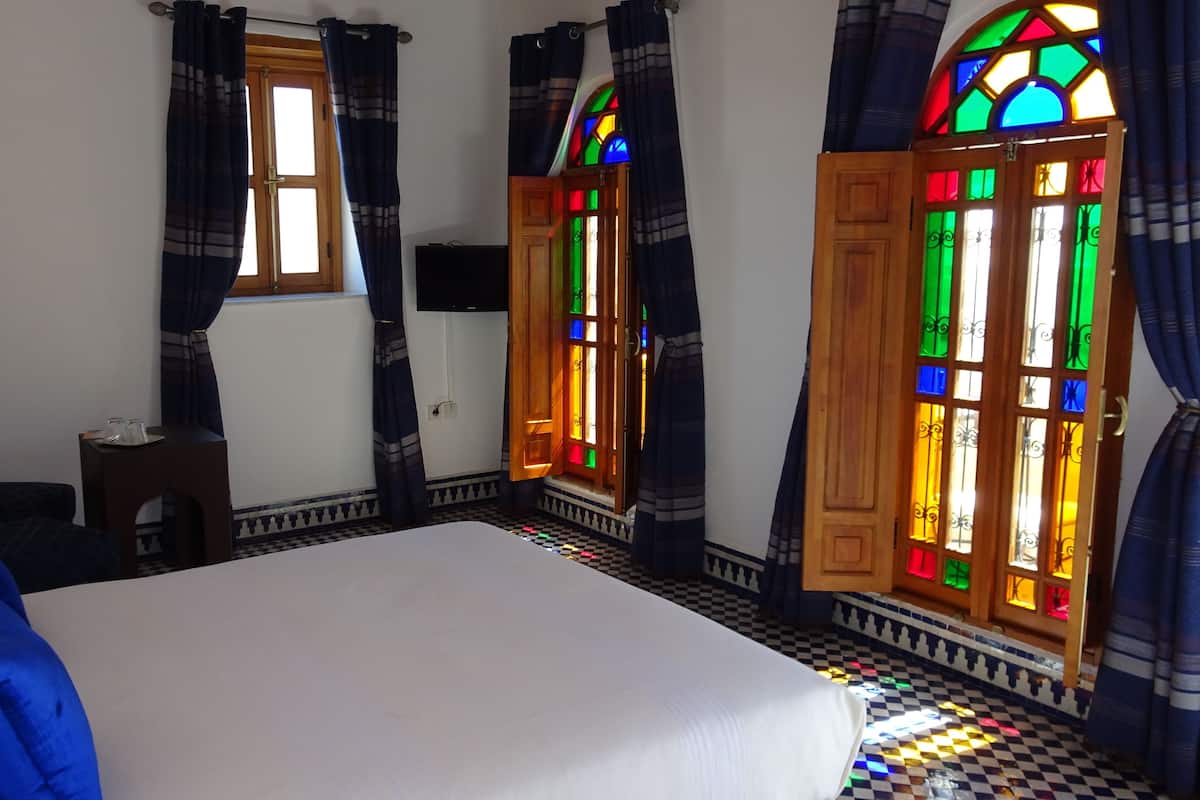 Una habitación con una cama, una mesita pequeña y dos ventanas con vidrieras de colores.