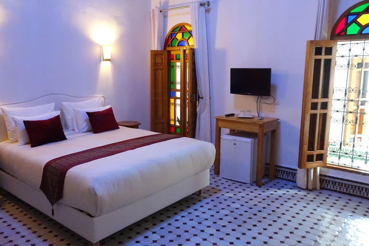 Una cama bien hecha con sábanas blancas y almohadas rojas, una mesita de noche, un televisor sobre un soporte de madera, un refrigerador y una ventana con un diseño de vitral.