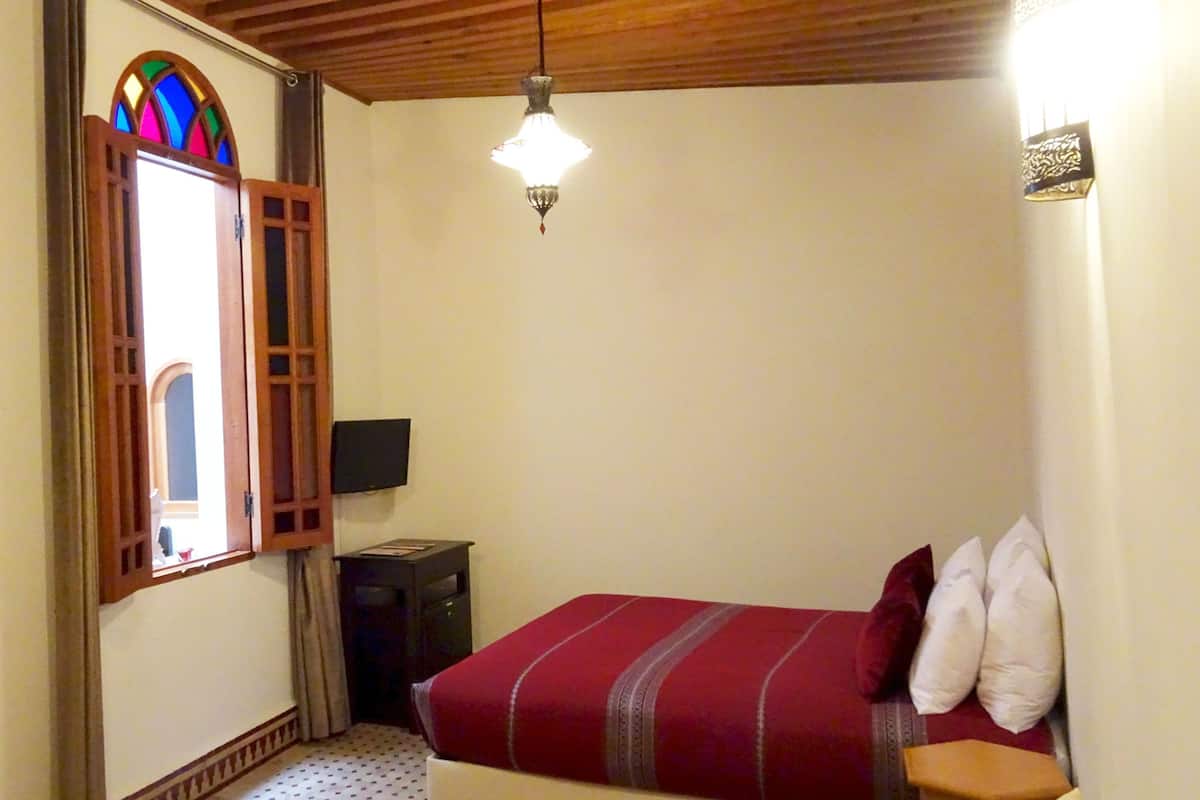 Un dormitorio con una cama, una puerta de madera, un ventanal con diseño de vitral, un televisor y una lámpara colgante.