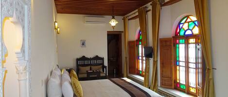 A bedroom with a bed, pillows, a nightstand, a wooden headboard, a wall-mounted air conditioner, a ceiling fan, and a window with stained glass.