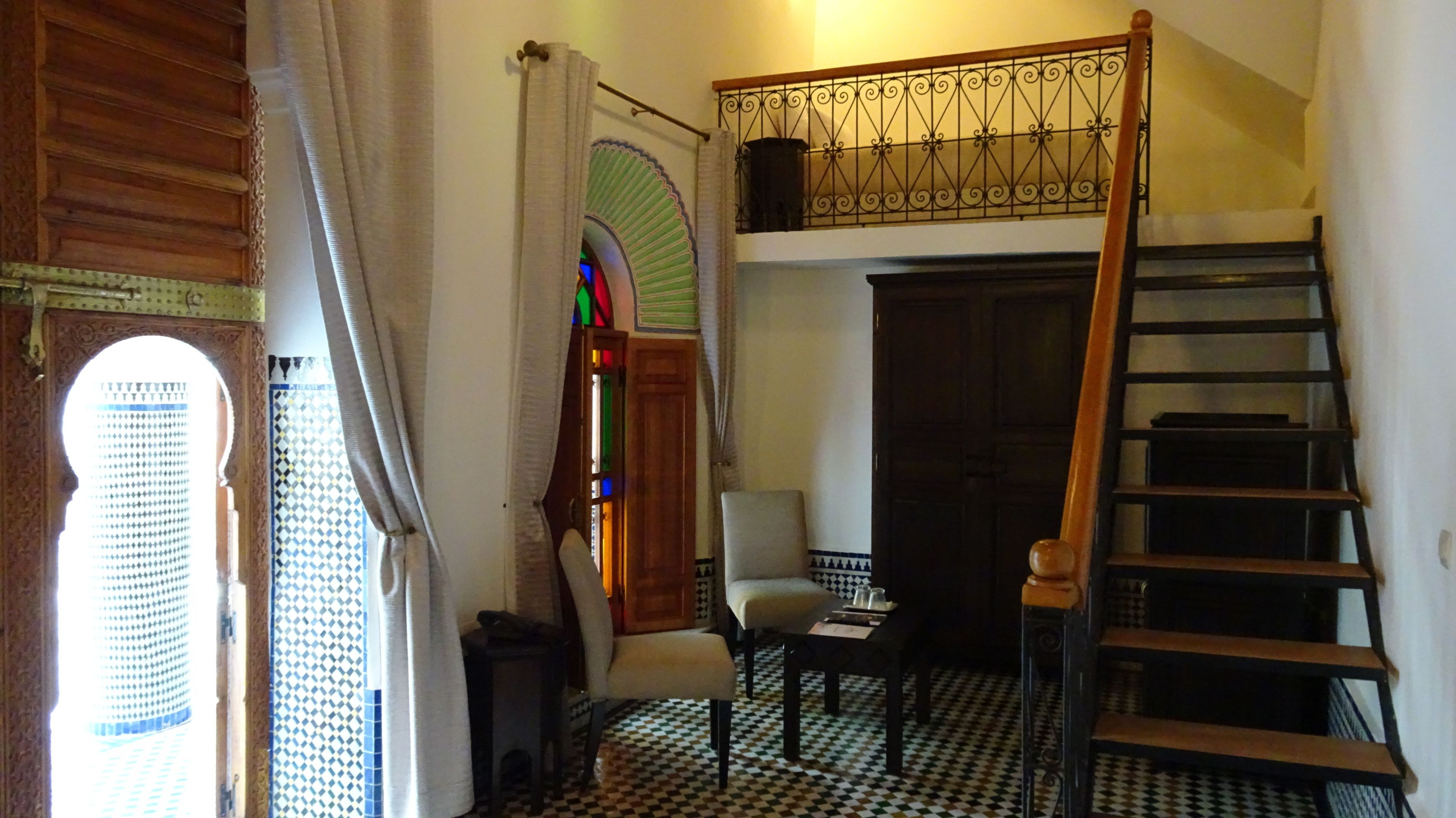 Una habitación con piso a cuadros, una escalera de madera, una silla, una mesita y una puerta con arco decorativo.