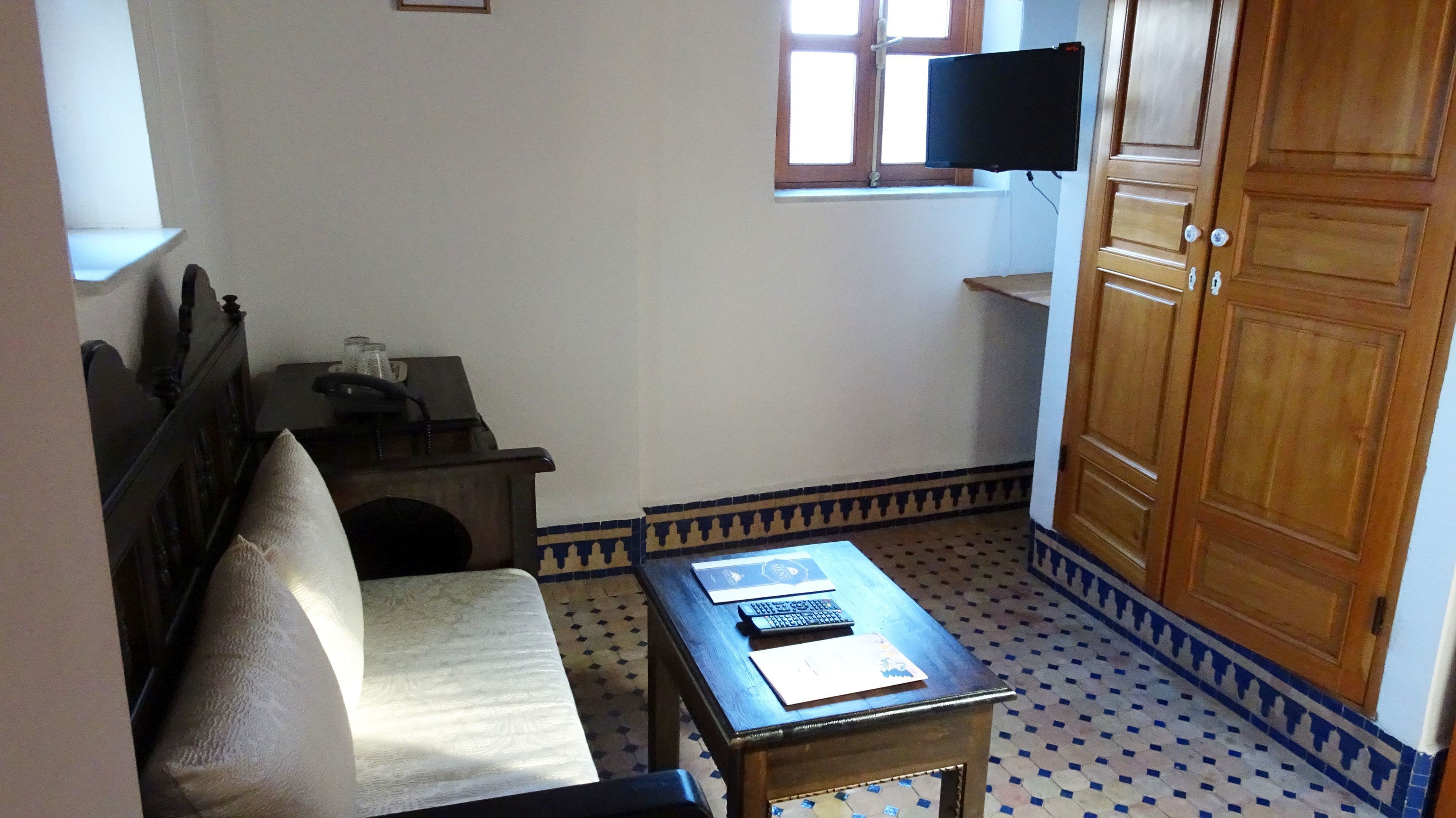 A small, compact room with a wooden wardrobe, a single bed, a small table with a keyboard, a television mounted on the wall, and a patterned floor.